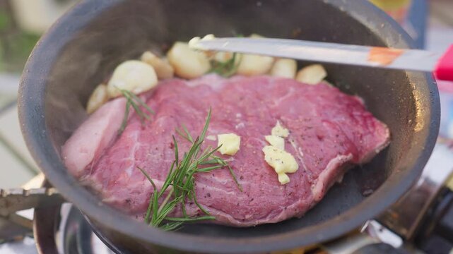 Raw beef steak seasoned with pepper sears in a hot pan with fresh rosemary, whole garlic cloves, and melting butter for a gourmet culinary preparation.