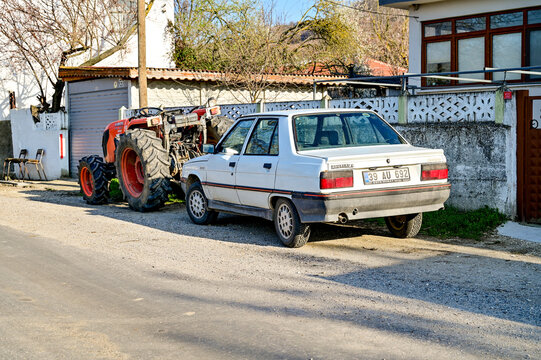 A white Turkish Renault 9 Broadway from the 1980s, now a classic car, and an orange tractor parked on a village road; rural life and cars in Kirklareli, Thrace, Turkey 28.03.2025