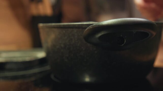 Low-angle close-up shows hand placing and lifting frying pan on glossy black induction cooktop, with visible handle grip, pan base contact, soft reflections, and warm modern kitchen background