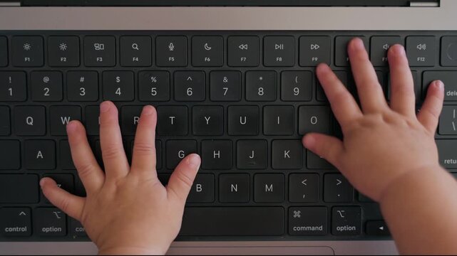 Funny cute top shot view on small baby hands typing on laptop computer keyboard. Quirky fun overhead shot of young toddler pressing on keys and buttons on modern laptop as if working in office or desk