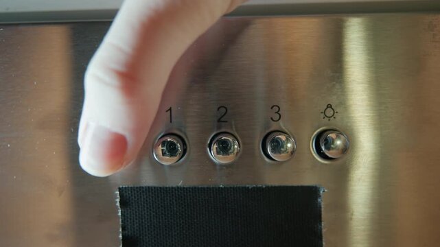 Close-up straight-on view of stainless steel appliance control panel with five round buttons aligned horizontally above black vent or display, fingertip pressing leftmost button, reflective brushed