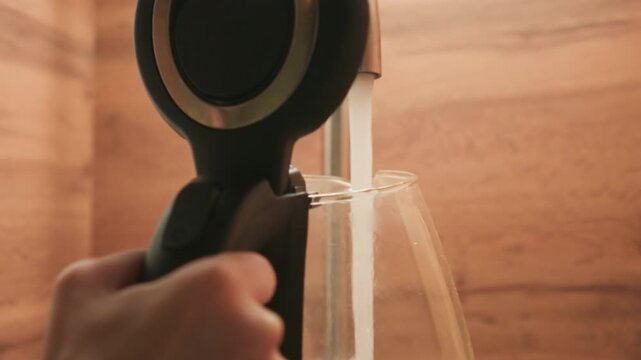 Hand holds black circular nozzle angled downward while clear water streams into glass beside warm wooden backsplash, showing steady motion, glossy reflections, and calm domestic routine under soft