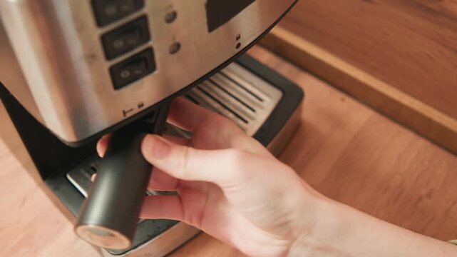 Closeup of hands guiding portafilter into stainless espresso machine on wooden counter, showing careful coffee preparation, manual rotation, secure locking, and warm cafe reflections