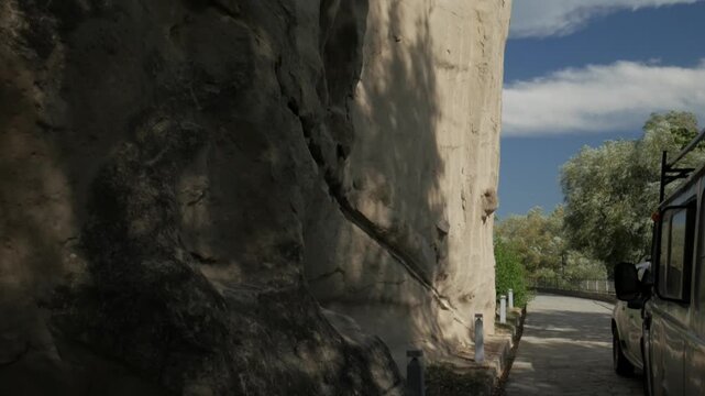 Handheld tilt down a massive sandstone cliff reveals a paved path and green trees in Uplistsikhe, Georgia. Bright blue sky contrasts with the rugged, ancient geological formation.