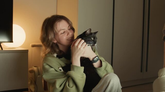 Woman seated indoors cradles small black dog close to her face, lifting and nuzzling pet with gentle kisses while supporting chest and head, creating warm domestic moment of care and affection