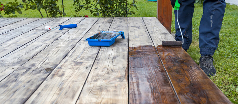 Person applies dark wood stain to weathered wooden deck planks