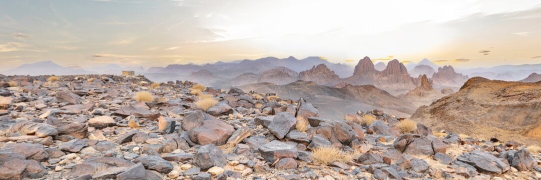 A panoramic landscape of the Hoggar Mountains in Assekrem, Tamanrasset, Algeria, showcasing rocky volcanic foreground terrain and sharp mountain spires under a warm brightening sunrise sky.