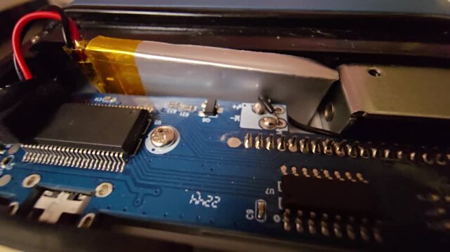 Close-up view of the internal components of a smart time-lock box, showing a printed circuit board (PCB), a rechargeable lithium-polymer battery, electronic microchips, and soldered wire connections 