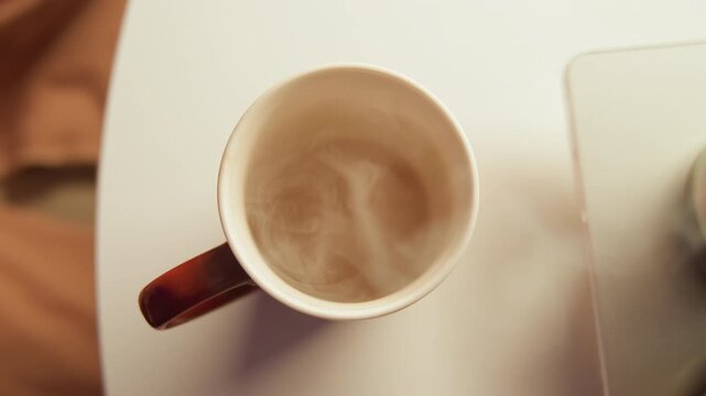 Top-down view shows hand grasping ceramic coffee cup by handle beside laptop corner on light desk, with rising steam, swirling surface, gentle tilt and calm morning workspace mood, with coffee, cup