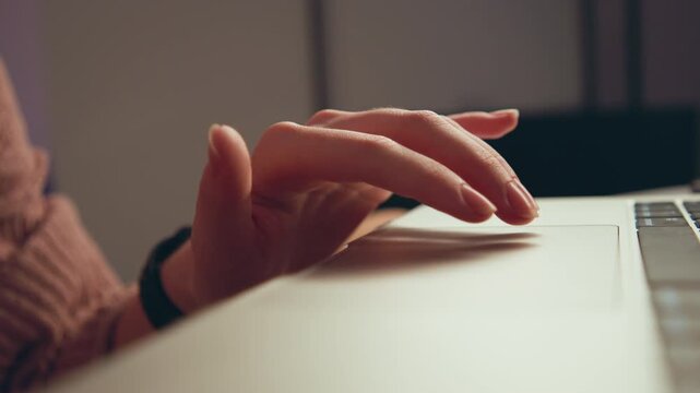 Diagonal close view of right hand sliding fingertips across laptop trackpad in warm indoor workspace, showing gentle scrolling motion, light contact, long sleeve forearm, calm focused productivity