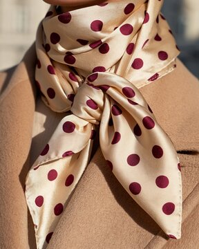Close-up photo of a fashion detail featuring a silk scarf with large burgundy polka dots tied elegantly around the neck of a person wearing a beige coat