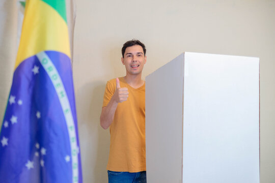 Jovem eleitor sorri e faz sinal de positivo ap&oacute;s votar, com a bandeira do Brasil desfocada em primeiro plano, nas elei&ccedil;&otilde;es presidenciais de outubro.