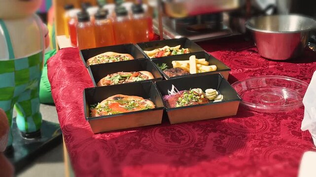 Food is arranged on a red tablecloth with boxes containing various dishes. Containers with drinks are visible, suggesting a vibrant market or food event