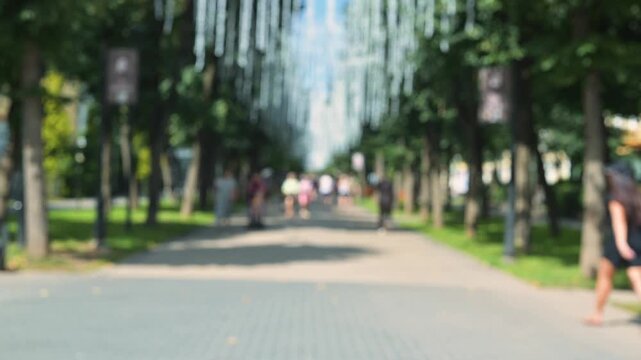 Groups of adults and children walk along a path in a park that features tall trees and hanging decorations, blurred