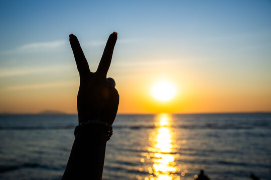 silhouetted hand making a peace sign against a glowing sunset over the ocean, with golden light