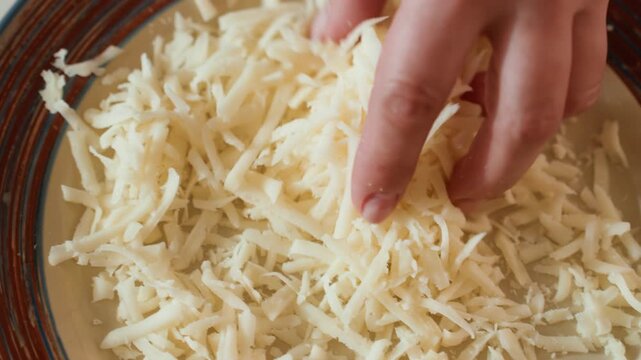 Close view shows hand sprinkling grated white cheese across shallow ceramic plate, shreds falling and gathering into soft textured pile, gentle kitchen preparation with natural light, fresh dairy