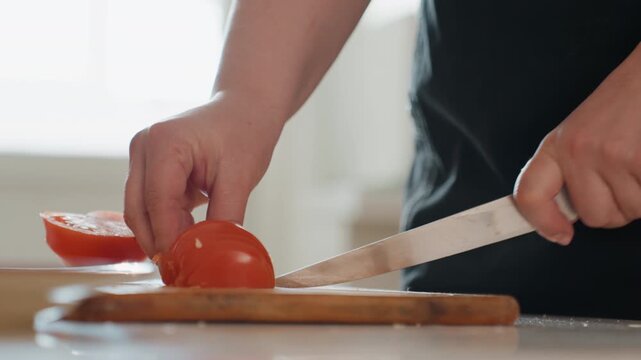 Close view of two adult hands slicing ripe red tomato on round wooden board, knife cutting through fruit while other hand steadies it, fresh slices, small pieces, soft natural daylight and clean