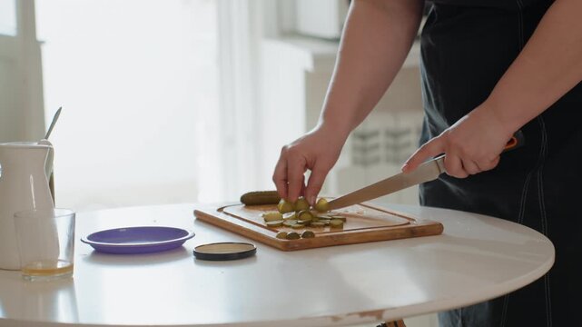 Hands cut green pickles on wooden cutting board beside round white table near bright window, with steady fingers, shining knife, and simple kitchen setting suggesting fresh food preparation