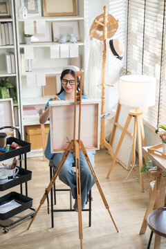 Young creative female artist sitting on a chair, painting on canvas with watercolors in a bright home art studio.