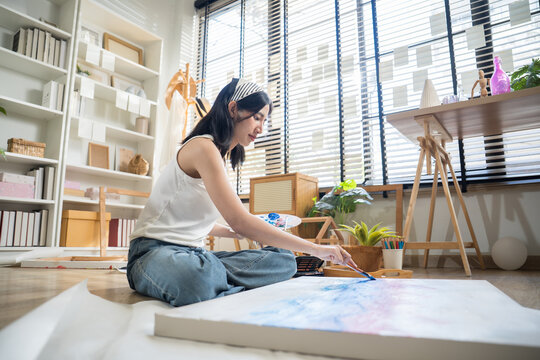 Creative tattooed young woman sitting cross-legged on the floor, mixing paint on a color palette to paint on paper in a bright home art studio.