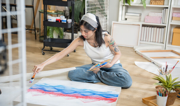 Creative tattooed young woman sitting cross-legged on the floor, mixing paint on a color palette to paint on paper in a bright home art studio.
