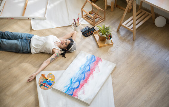 Creative tattooed woman lying on wooden floor, resting peacefully next to her completed canvas artwork with satisfaction.