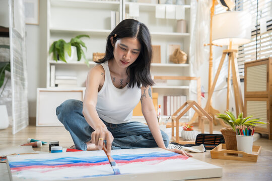 Creative tattooed young woman sitting cross-legged on the floor, mixing paint on a color palette to paint on paper in a bright home art studio.