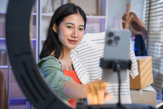 Young woman holding a shirt on a hanger while live streaming to sell fashion clothing using a smartphone on a tripod ring light.