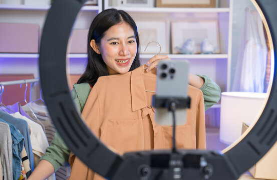 SME retail store owner smile enjoy selling show live sale online by using her mobile phone to record videos while showcasing her clothing designs. Asian female fashion designer concept