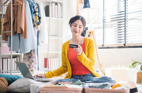Happy woman sitting on the floor with her luggage, holding a credit card and using a laptop to make an online hotel reservation.