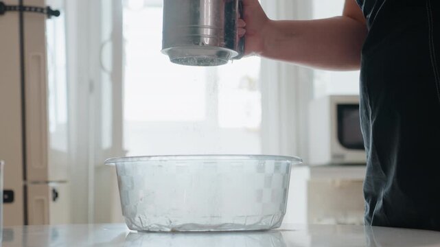 Right hand in dark short sleeve shirt holds metal sifter above textured glass mixing bowl on countertop, showing kitchen prep for baking with clean bright daylight, with sifter, bowl, kitchen, hand