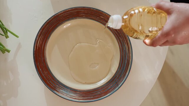 Top-down view shows right hand tilting plastic bottle to pour clear cooking oil into center of white ceramic plate with brown patterned rim, creating glossy pool during kitchen prep