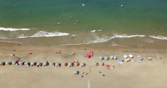 Aerial zenithal view of a sandy beach with colorful umbrellas along the shore, people swimming and relaxing in the clear turquoise sea, gentle waves breaking on the golden sand in summer.
