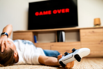 Frustrated woman with game controller in hand lying on floor after losing video game with “Game Over” displayed on TV © Igor Barilo