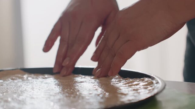 Close-up of two hands pressing and smoothing thin batter or dough inside round metal baking pan, with gentle fingertip spreading, soft daylight, subtle surface dimpling, ripples, and calm home