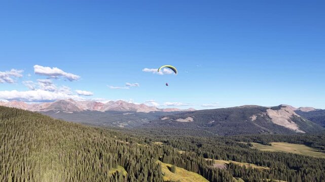 Aerial of paragliding over San Juan National Forest in south Colorado in summer with vast coniferous forests and red colored mountain ranges under a blue sky with scattered clouds