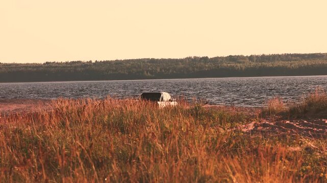 Pickup truck parked on a sandy lake shore, partially hidden behind tall golden grass, distant tree lined opposite shore, warm evening sunset light, calm water, remote quiet scenic landscape backdrop
