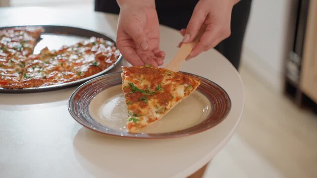 Adult hands use spatula to lift slice from round baked dish on black tray and place onto patterned ceramic plate, with melted cheese stretching slightly, browned crust, herbs, and warm indoor light