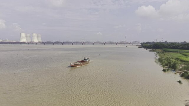Drone aerial footage of the Padma River, with Hardinge Bridge and Rooppur Nuclear Power Plant cooling towers far away, while loaded cargo boats move toward the camera across the river.