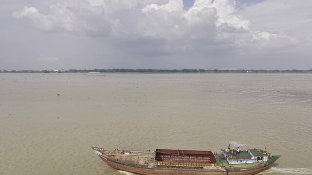 Drone footage taking a half orbit around a cargo boat on the wide Padma River in Bangladesh, showing the moving vessel, its wake, green riverbank,  and cloudy sky