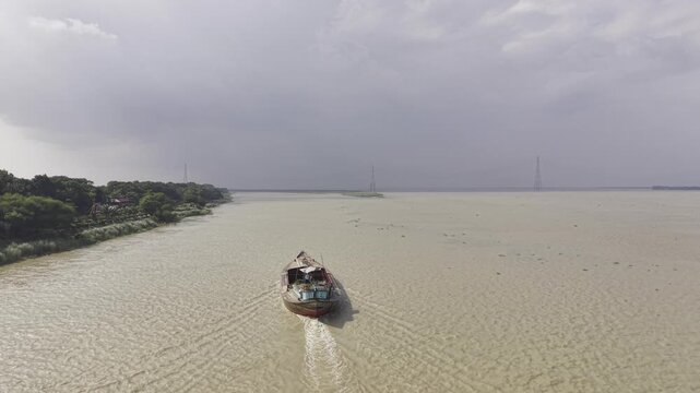 Aerial drone footage of a cargo boat approaching across the wide Padma River in Bangladesh, leaving a visible wake, with green riverbank vegetation, transmission towers and large clouds.

