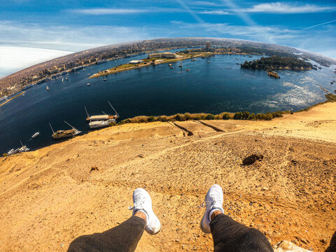 View of the Nile River from a high sandy hill with a person's feet visible in the foreground, featuring boats on the water, Aswan, Aswan Governorate, Egypt.