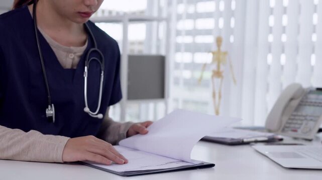 Asian female physician reviewing patient medical documents at modern clinic desk with skeleton model