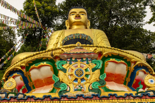 View of the golden Buddha statue adorned with intricate colorful details and surrounded by vibrant prayer flags and lush green trees in Pokhara, Gandaki Province, Nepal.