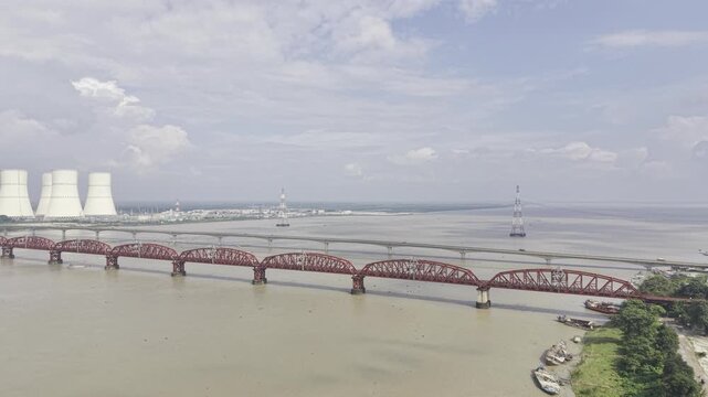 Aerial view of the Padma River with Lalon Shah Bridge, Rooppur Nuclear Power Plant cooling towers, power infrastructure and cargo boats under a blue sky with dramatic white clouds in Bangladesh.