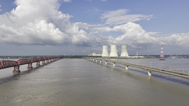 Slowly ascending drone footage over the Padma River between Hardinge Bridge and Lalon Shah Bridge, revealing Rooppur Nuclear Power Plant cooling towers and Green City Rooppur buildings.