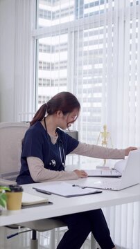 Young Asian female doctor reviewing medical documents on laptop in modern clinic office with city view