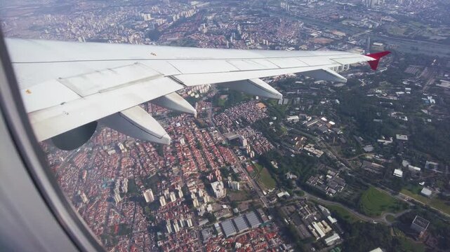 Aerial view through a passenger window showing an airplane wing with a red tip flying over the vast urban cityscape and dense architecture of Sao Paulo, during a daytime commercial flight.