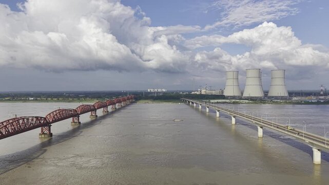 Forward-moving drone footage between Hardinge Bridge and Lalon Shah Bridge over the Padma River, showing Rooppur Nuclear Power Plant cooling towers, Green City Rooppur buildings, blue sky and clouds.