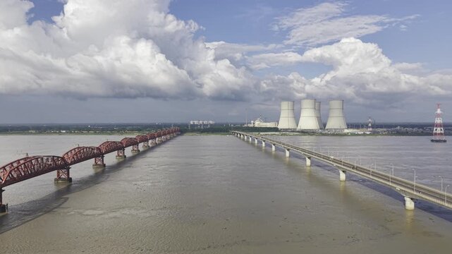 Forward drone footage between Hardinge Bridge and Lalon Shah Bridge over the Padma River, capturing Rooppur Nuclear Power Plant cooling towers, Green City Rooppur buildings, blue sky and clouds.
